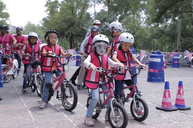 Kinder fahren mit Fahrrädern auf einer Straße mit Verkehrskegeln, tragen Helme und einige haben Gesichtsfarbe, vor einem Hintergrund aus Bäumen und einem klaren blauen Himmel.
