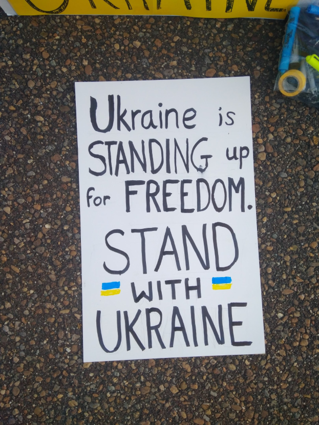 Weißer Zettel mit dem Text "Ukraine steht für Freiheit, steht mit Ukraine" auf dem Boden vor einer gelben Fahne, mit einer Plastikhülle rechts daneben.