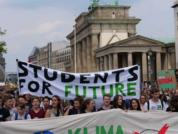 Gruppe von Studenten marschiert in Berlin mit buntem "Students for Future"-Schild gegen Gebäude, Bäume und Himmel.