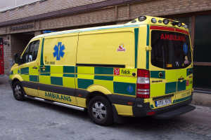 Ambulanz vor einem Backsteingebäude mit einem Feuerlöscher links daneben geparkt, auf einer Straße mit lesbarem Text auf dem Gebäude im Hintergrund.