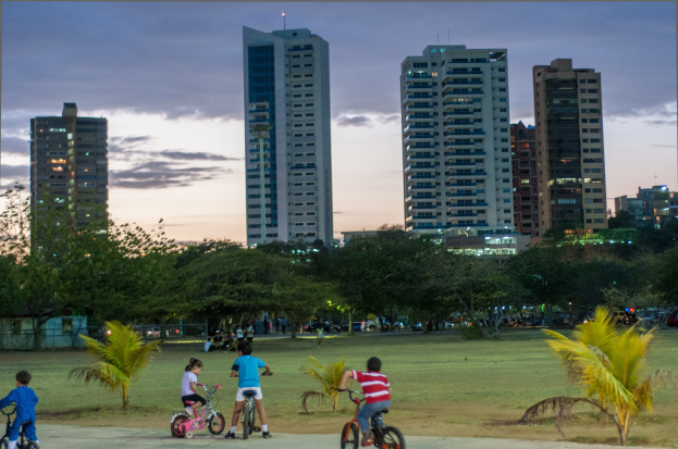 Kinder auf Fahrrädern in einem Park bei Sonnenuntergang mit saftigem Grün und Bäumen, hohe Wolkenkratzer vor einem Himmel aus Orangen, Pinks und Purpurs.
