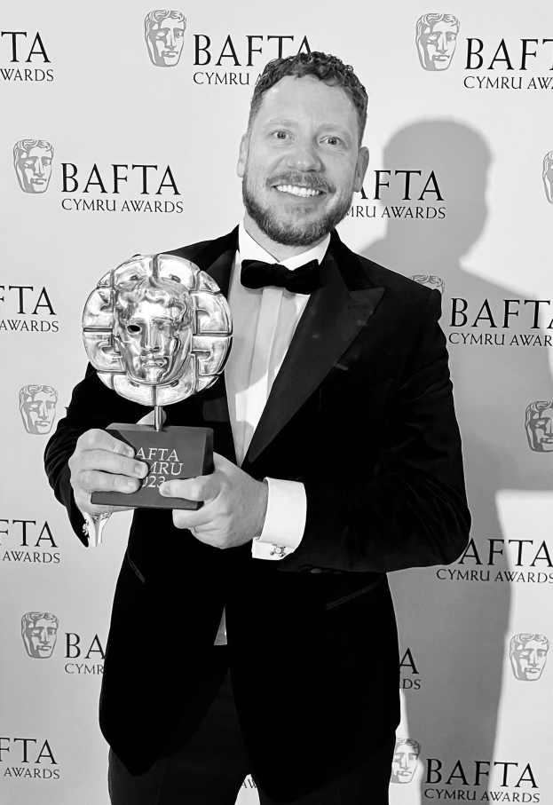 Ein Mann im Smoking, der eine BAFTA-Auszeichnung hält und lächelt, mit einem Hintergrundplakat, auf dem "BAFTA Awards 2018" steht.
