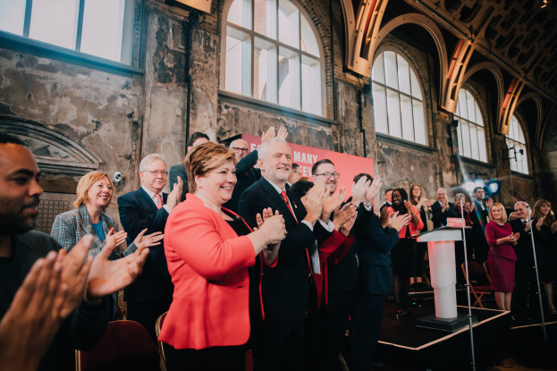 Eine Gruppe von Menschen, die vor einem Publikum stehen und applaudieren, näher an einem Podium mit einem Mikrofon, einem Banner mit der Aufschrift "Covid-19 New York City Mayor Bill de Blasio" und Hintergrundelementen wie Stühlen, einer Wand und Fenstern.