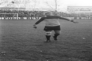 Professioneller Fußballspieler in blauer Uniform auf einem Feld mit Publikum, Tor und Bäumen im Hintergrund.