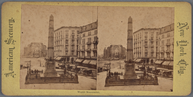 Altes Schwarz-Weiß-Foto einer Stadtstraße mit einem zentralen Denkmal, flankiert von Gebäuden, Straßenmöbeln, geparkten und fahrenden Fahrzeugen, Fußgängern auf dem Gehweg und einem sichtbaren Himmel.
