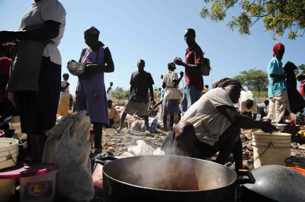Eine Gruppe von Menschen bereitet Essen im Freien in einer ländlichen Gegend zu, mit einem großen Topf, Eimern und Abdeckungen auf dem Boden, unter einem klaren blauen Himmel.