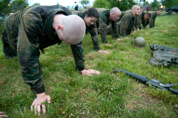 Eine Gruppe von Männern in Militäruniformen, die Liegestütze auf dem Gras machen, mit einer Waffe, einem Helm und anderen Gegenständen drumherum, mit Bäumen, Lichtern, Fahrzeugen und einem klaren blauen Himmel im Hintergrund.