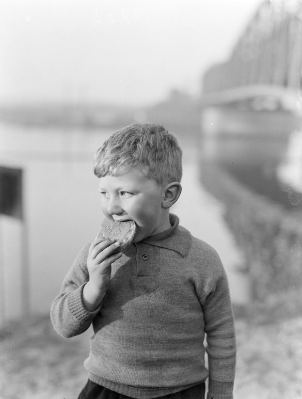 Ein junger Junge in einem alten Schwarz-Weiß-Foto hält ein Stück Brot in der Hand, während er mit einem unscharfen Hintergrund steht.
