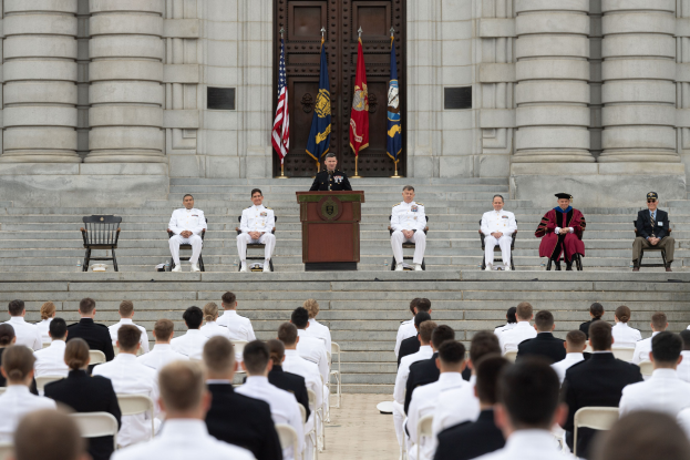 Gruppe von Menschen, die auf Stühlen sitzen und auf ein Gebäude mit Säulen und einer Tür schauen, mit einem Podium und einem Mikrofon in der Mitte und Fahnen im Hintergrund, was auf eine Abschlussfeier hindeutet.