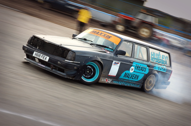 Ein Auto fährt auf einer Rennstrecke mit Rauch hinter sich, umgeben von anderen Fahrzeugen und Menschen, mit einem unscharfen Hintergrund, der hohe Geschwindigkeit andeutet.