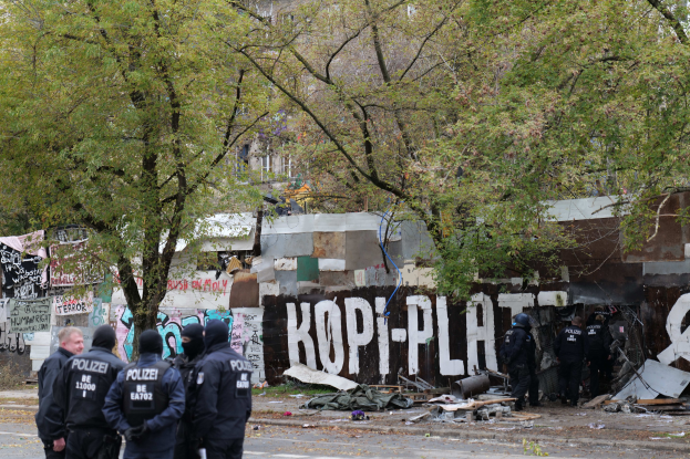Polizisten in schwarzen Uniformen und Helmen stehen vor einem mit Graffiti bedeckten, baufälligen Gebäude mit verstreuten Trümmern und Bäumen im Hintergrund.