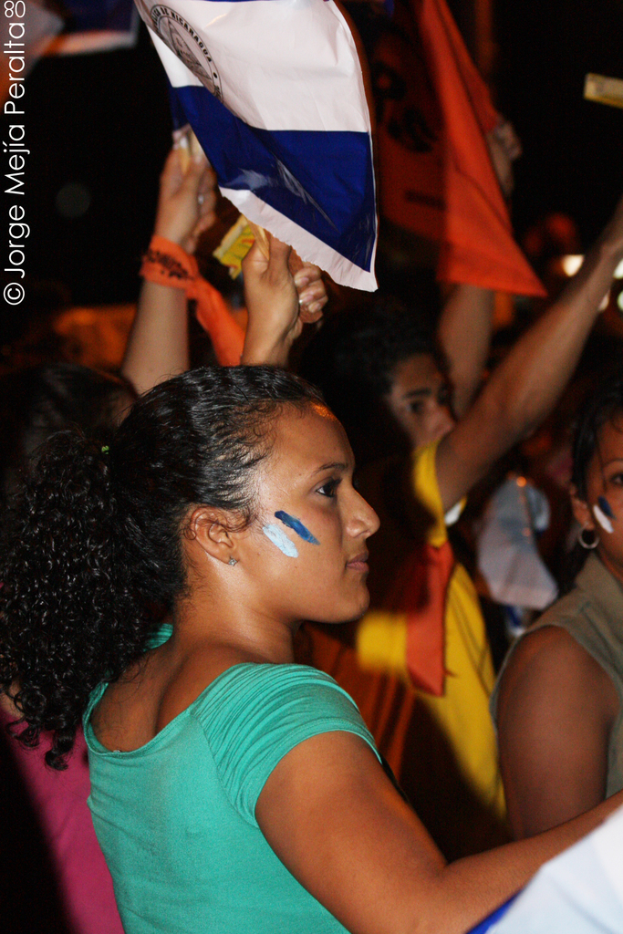 Eine Frau in einem grünen T-Shirt steht bei einer Demonstration und hölt ein weißes Schild, mit vielen anderen Menschen dahinter.