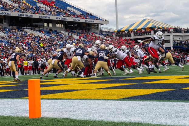 Fußballspieler mit Helmen konkurrieren auf einem Rasenfeld, während Zuschauer im Hintergrund unter einem bewölkten Himmel sitzen, wobei ein Zelt, ein Pfahl und ein Gegenstand auf dem Boden sichtbar sind.