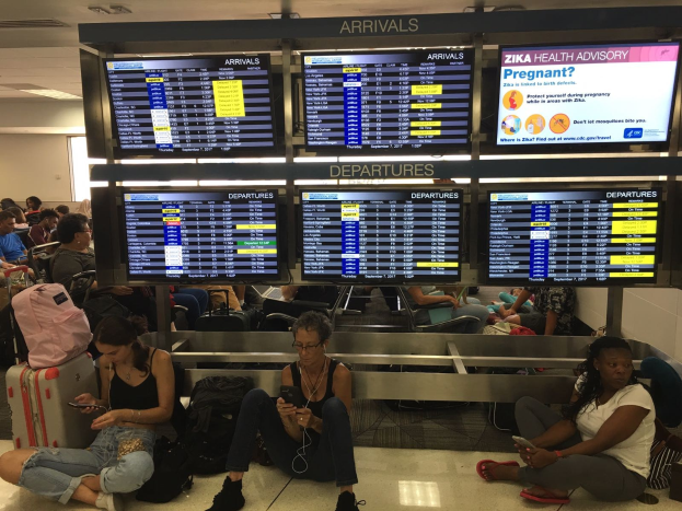 Eine Gruppe von Menschen sitzt auf dem Boden eines Flughafens, einige halten Handys, mit verstreuten Taschen und Bildschirmen mit Zika-Virus-Information im Hintergrund.