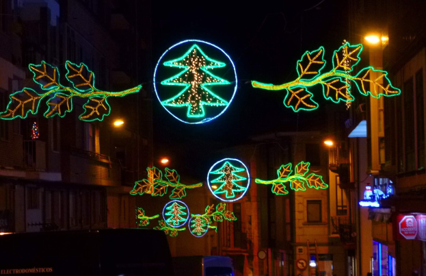 Eine städtische Straße mit festlichen Weihnachtslichtern, die Blätter und andere Schmuckelemente tragen, mit Fahrzeugen, die an Gebäuden auf beiden Seiten vorbeifahren.