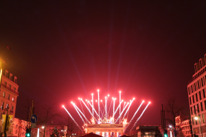 Eine nächtliche Stadtstraße mit Gebäuden, Bäumen, Laternen, Verkehrsampeln, Schildern und Menschen, mit Feuerwerk, das den Himmel erhellt.