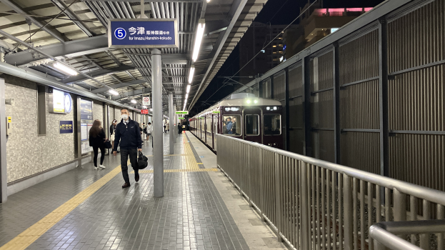 Ein Zug fährt auf einem Bahnsteig ein, mit Menschen in der Nähe, einem Geländer auf der rechten Seite, Gebäuden im Hintergrund, Bahnsteiglampen und einer Anzeigetafel.