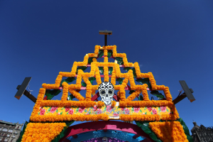 Ein Día de los Muertos-Bogen, geschmückt mit Blumen und einem Schädel, steht in Mexiko-Stadt, eingerahmt von Gebäuden unter einem klaren blauen Himmel.