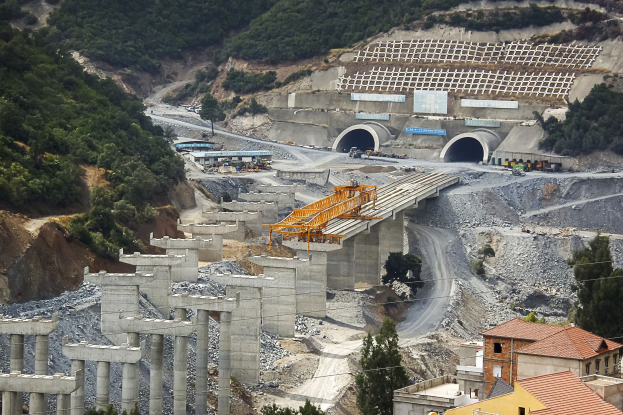 Großer Tunnel an einer Baustelle umgeben von Bäumen, Gebäuden, Fahrzeugen, Säulen, Felsen und einer Straße.
