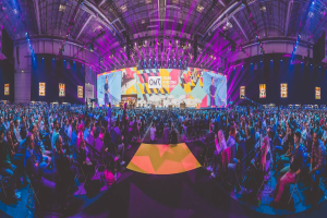 Eine große Menschenmenge sitzt auf Stühlen, die einer hell erleuchteten Bühne zugewandt sind, mit mehreren stehenden Individuen, wahrscheinlich auf einem Online Marketing Rockstars-Event, umgeben von Bildschirmen.