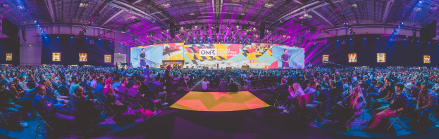 Eine große Menschenmenge sitzt auf Stühlen, die einer hell erleuchteten Bühne zugewandt sind, mit mehreren stehenden Individuen, wahrscheinlich auf einem Online Marketing Rockstars-Event, umgeben von Bildschirmen.