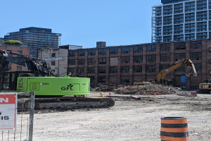 Eine Baustelle mit einem großen grünen Bagger in der Mitte, umgeben von einem Metallzaun mit einem Schild an einem Pfahl, Maschinen, einem Eimer und einem Verkehrskegel im Vordergrund und Gebäuden, Bäumen und Himmel im Hintergrund.