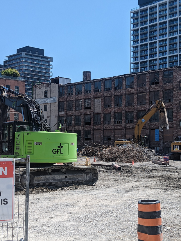 Eine Baustelle mit einem großen grünen Bagger in der Mitte, umgeben von einem Metallzaun mit einem Schild an einem Pfahl, Maschinen, einem Eimer und einem Verkehrskegel im Vordergrund und Gebäuden, Bäumen und Himmel im Hintergrund.