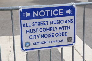 Anschlagtafel mit einem Schild, das die Einhaltung städtischer Lärmschutzvorschriften für Straßenmusiker verlangt, an einem Zaun befestigt.