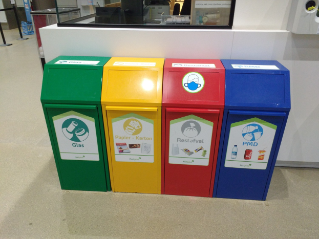 Vier bunte Mülltonnen in einer Reihe auf dem Boden, mit einer Arbeitsplatte und Gegenständen im Hintergrund und einer Person auf der linken Seite.