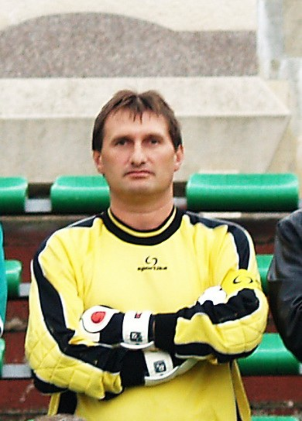 Ein Torwart in gelbem Trikot steht mit konzentrierter Miene neben einem Mann in schwarzer Jacke, mit grünen Stühlen und einer Wand im Hintergrund.