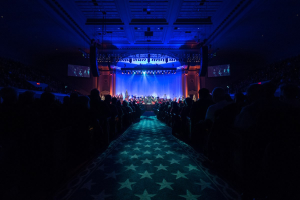 Ein großer Saal mit Menschen auf Stühlen, eine Bühne mit Musikern und eine Decke mit Lichtern und Fernsehern, was auf ein Konzertsaal während der Nationalkonvention 2019 hinweist.
