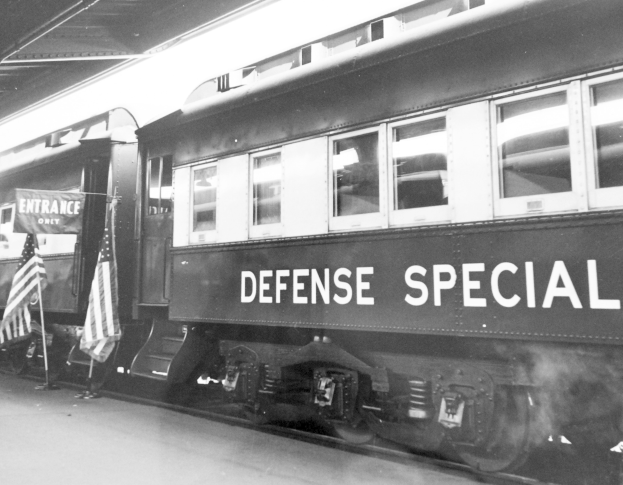 Schwarz-weiß-Foto eines Zuges mit der Aufschrift "Defense Special" auf den Gleisen geparkt mit zwei Fahnen links und einer Decke oben.