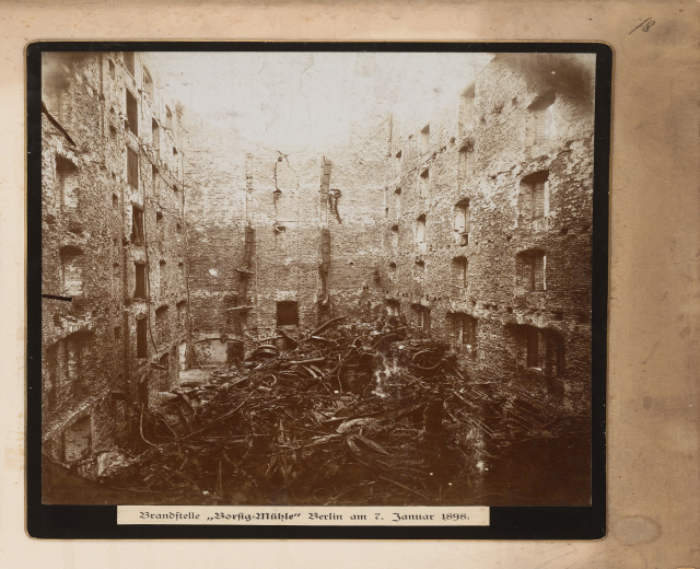 Ein Schwarz-Weiß-Foto eines zerstörten städtischen Gebäudes, das von einem Photo-Rahmen umgeben ist, mit Schutt und zerbrochenen Fenstern und Text am unteren Rand.