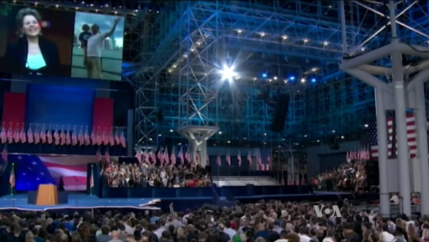 Eine große Menschenmenge sitzt vor einer Bühne mit einem Podium, einem Mikrofon, Flaggen, einem Bildschirm und Beleuchtungsausrüstung und wartet auf eine Debatte.