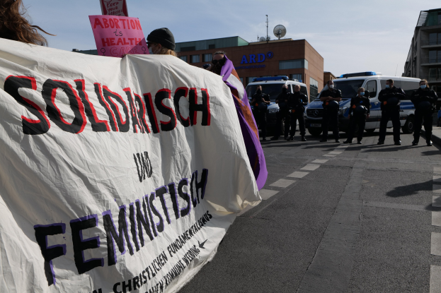 Eine Gruppe von Menschen marschiert auf einer Straße und trägt ein Banner mit der Aufschrift "Solidarität und Feminismus", mit parkenden Fahrzeugen, Gebäuden, einer Schüsselantenne und einem klaren blauen Himmel im Hintergrund.