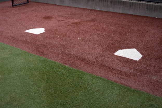 Baseballfeld mit Kunststoffrasen, umgeben von einem Zaun, mit Home Plate in der Mitte und einer Wand im Hintergrund.
