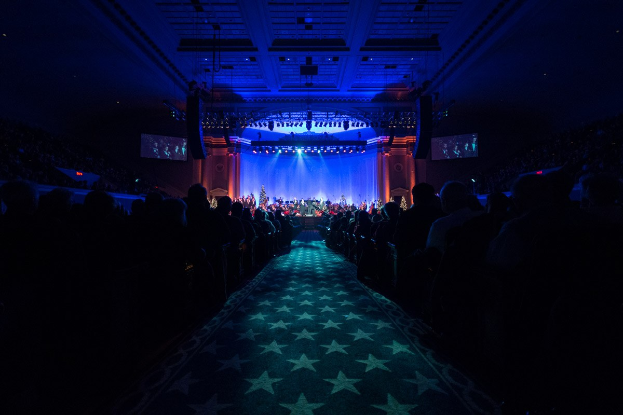 Ein großer Saal mit sitzenden Zuschauern, eine Bühne mit Musikern und Deckenbeleuchtung und -bildschirme, was auf einen Konzertsaal während der Nationalkonvention 2019 hindeutet.