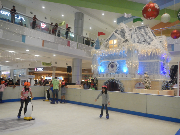 Eine Gruppe von Menschen, die auf einer Eisbahn in einem Einkaufszentrum Schlittschuhlaufen, umgeben von einem hausähnlichen Aufbau, der mit Lichtern und festlichen Dekorationen geschmückt ist, mit Menschen in der Nähe und Lichtern an der Decke.