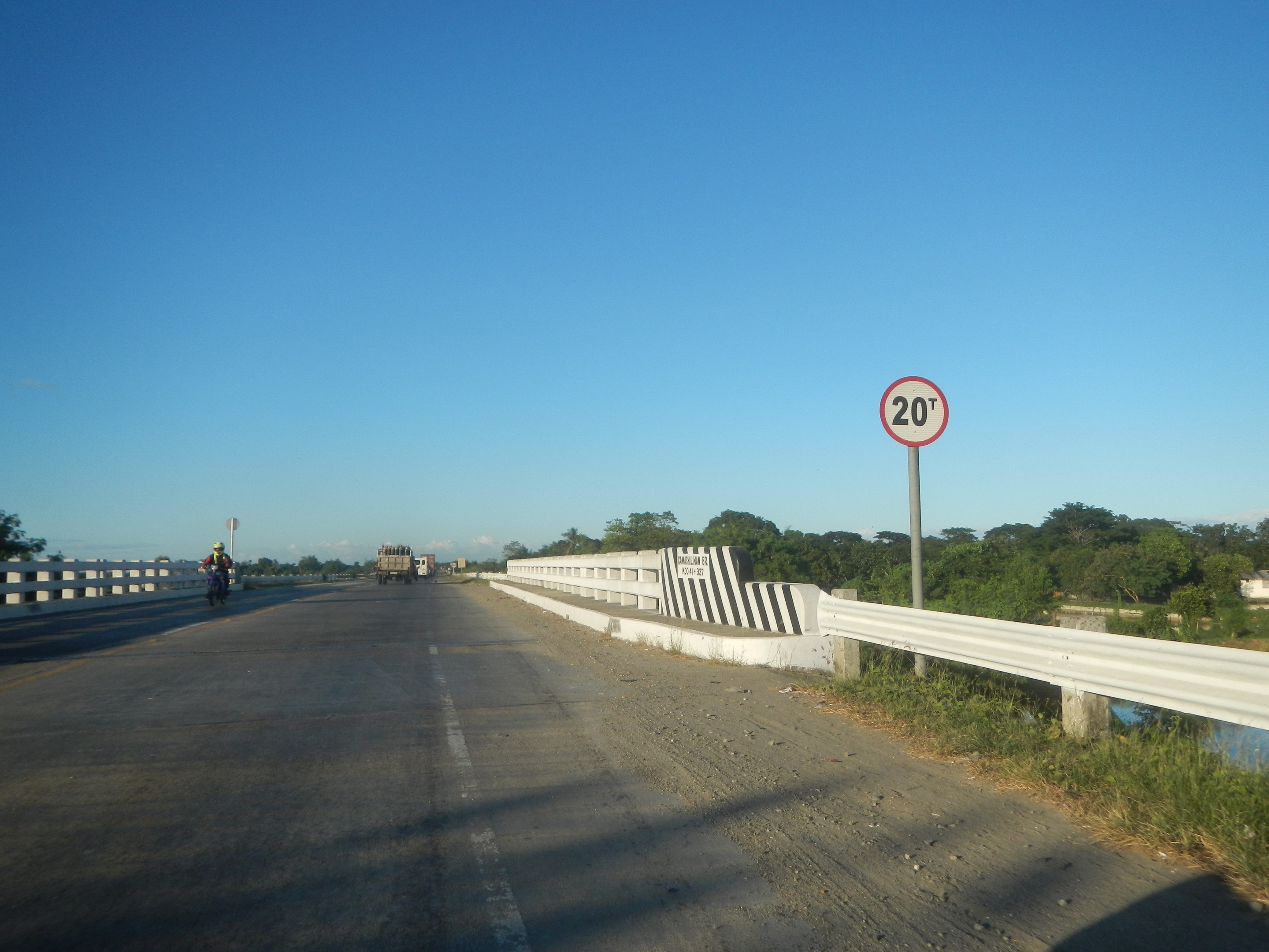 Fahrbahn mit fahrenden Fahrzeugen, ein Tempolimit-Schild, Geländer, Gras, Bäume und einen klaren blauen Himmel.