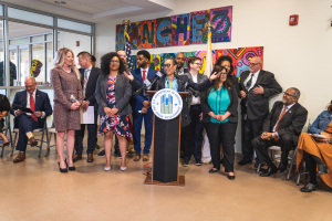 Gruppe von Menschen in einem Raum mit einem Podium, Mikrofon und einer Tafel mit der Aufschrift "Sancho-Stadtrat tritt dem Stadtrat bei", umgeben von Flaggen, Stühlen und Deckenleuchten.