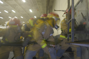 Feuerwehrleute in Helmen und Schutzausrüstung arbeiten an einem Flugzeug, halten verschiedene Gegenstände und haben eine Leiter auf der rechten Seite, unter Deckenlampen, wahrscheinlich während eines Brandes.