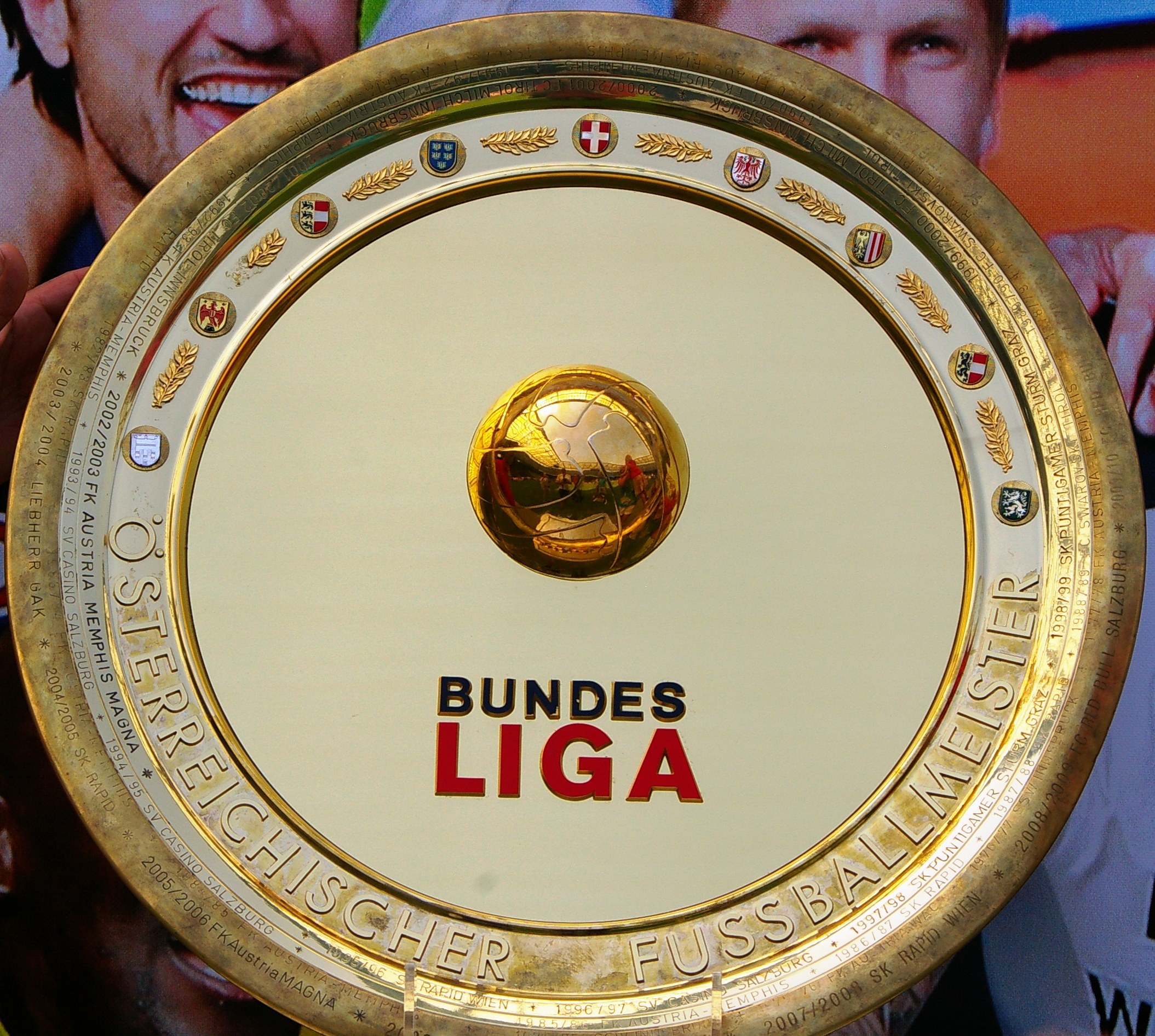Ein Mann mit einem goldenen Schild mit der Aufschrift 'Bundesliga', im Hintergrund eine Fahne mit drei Personen.