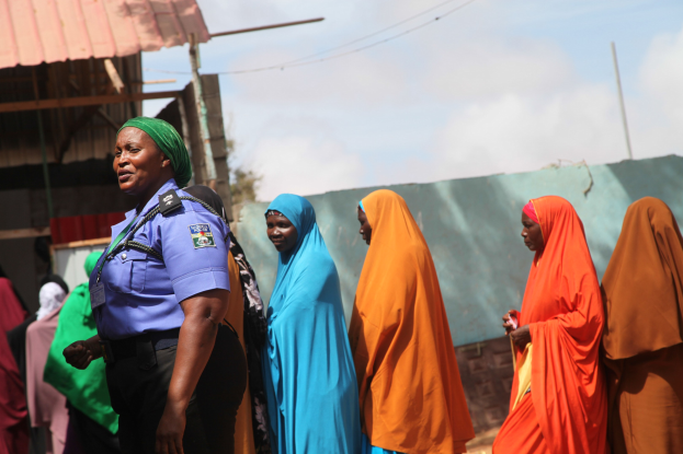 Gruppe von Frauen neben einem Gebäude mit einem Polizeibeamten im Vordergrund, Schuppen, Pfähle, Drähte, Bäume und einen klaren blauen Himmel im Hintergrund.