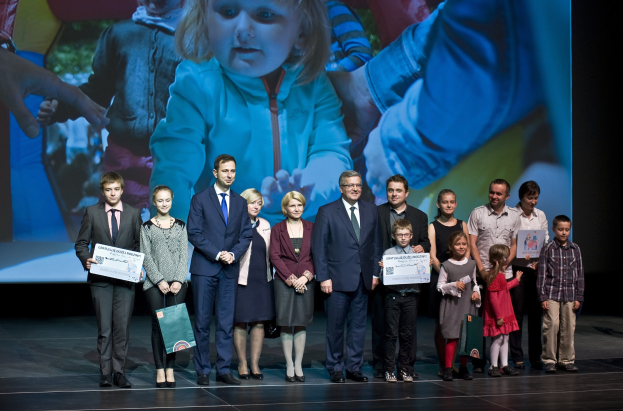 Gruppe von Menschen auf einer Bühne mit Taschen und Urkunden, im Hintergrund ein Bildschirm mit einem Bild von einem Kind und anderen