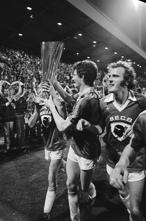 Schwarzes und weißes Foto von Fußballspielern, die einen Pokal auf einem Feld halten und ihren Sieg in der FA-Cup-Finalbegegnung feiern, während Zuschauer hinter einem Zaun zusehen.