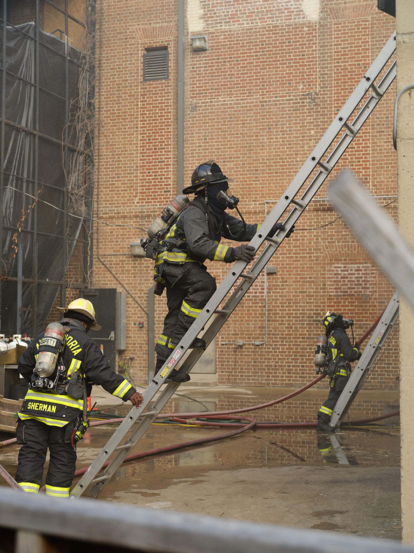 Feuerwehrleute in Helmen und Ausrüstung klettern an einer Leiter vor einem Backsteingebäude mit Rohren auf dem Boden und einer Metallstange am Boden, mit einem weiteren Gebäude mit Fenstern und einem Netz im Hintergrund.