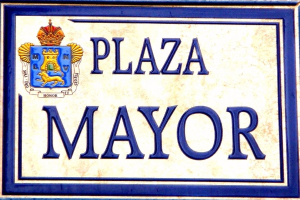 Rechteckige weiße Plakette mit schwarzer Schrift "Plaza Mayor" in fetter Schrift, blauem Rand und einem zentralen gekrönten Wappen-Logo an einer Gebäudefassade.