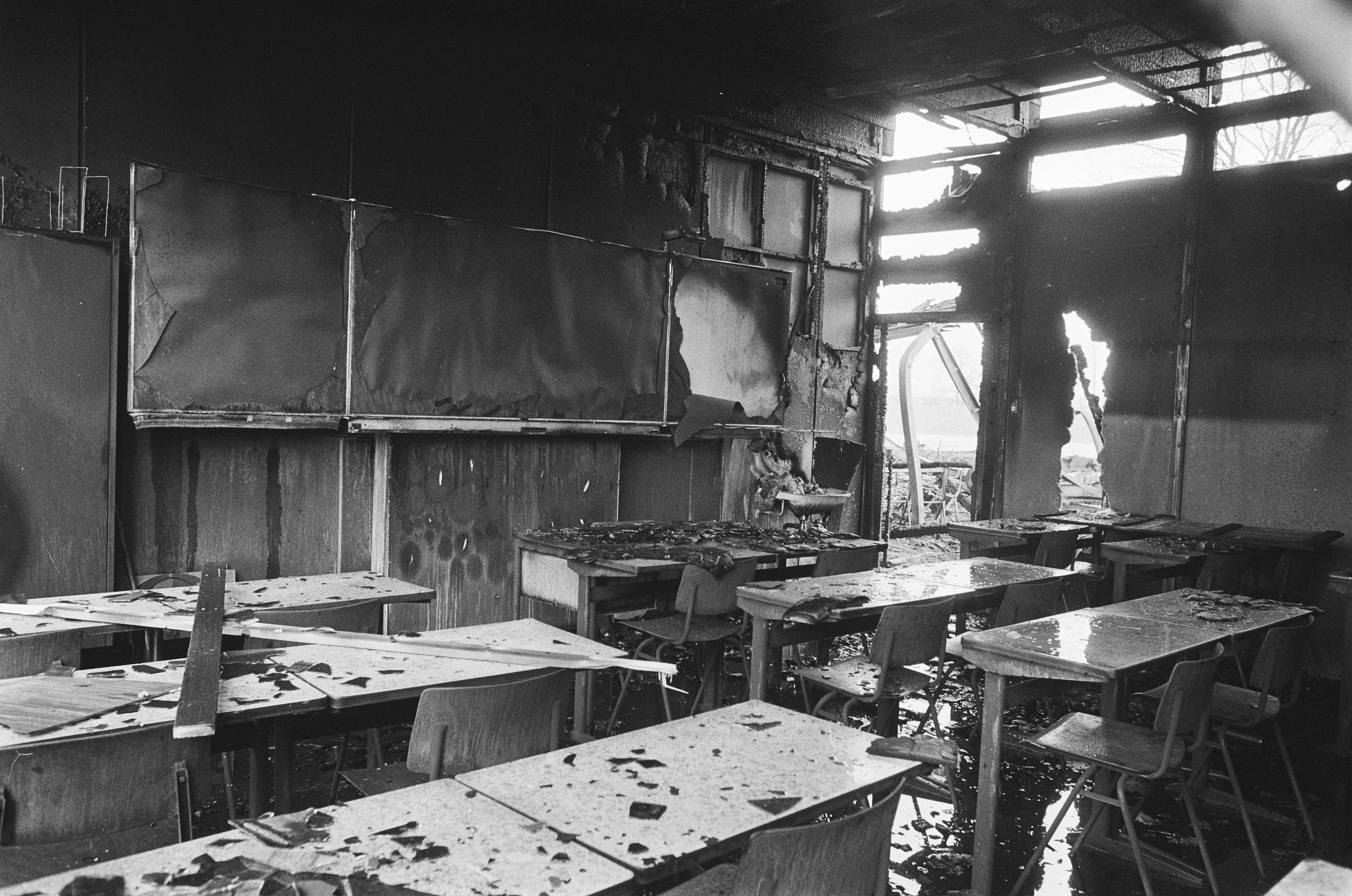 Schwarz-weißes Foto eines Klassenzimmers mit verkohlten Wänden, zerbrochenen Fenstern und verstreuten Tischen und Stühlen, das eine trostlose und verlassene Atmosphäre vermittelt.